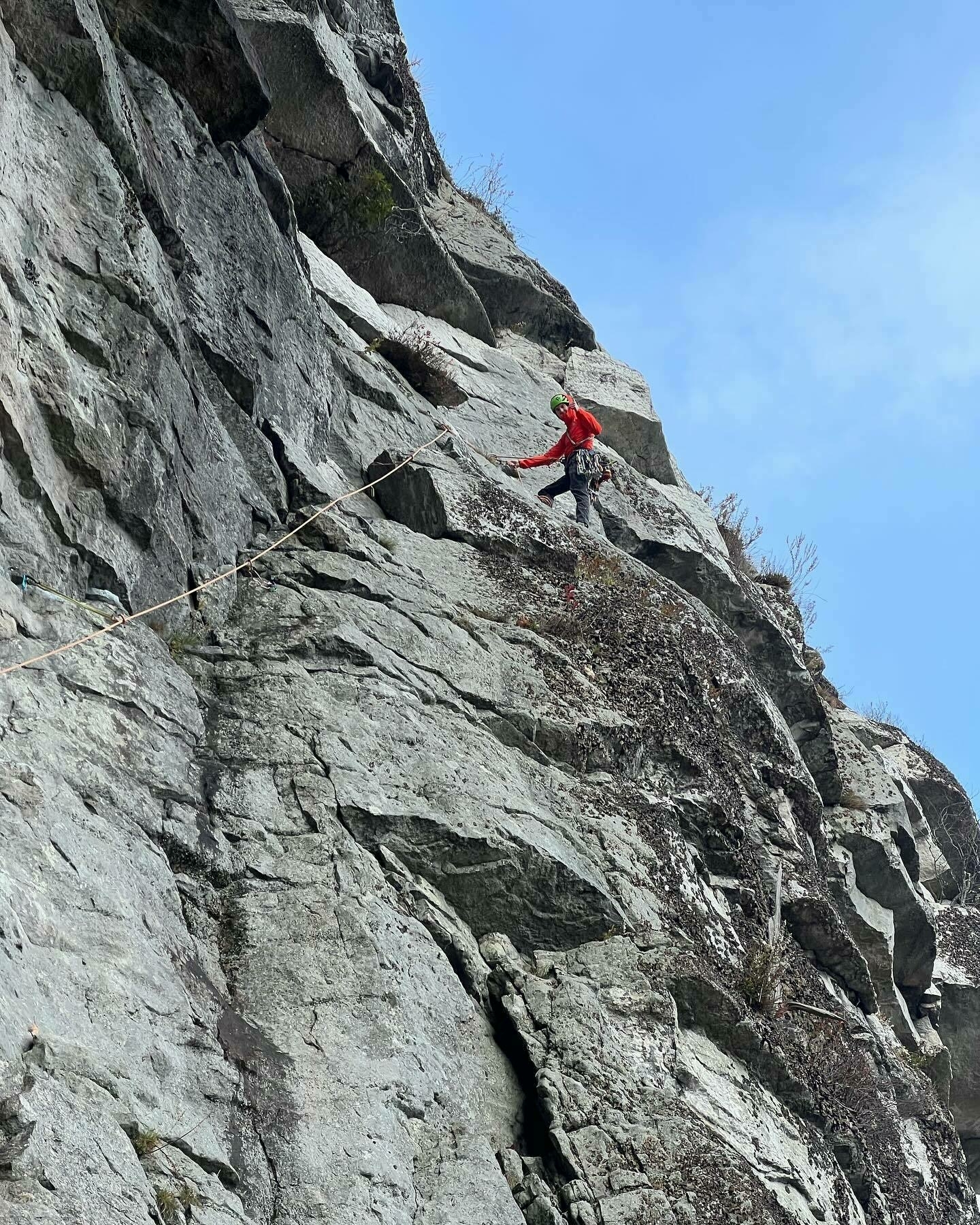 A climber in red gear ascends a steep, rocky cliffside against a clear sky, secured by ropes on a bright day.