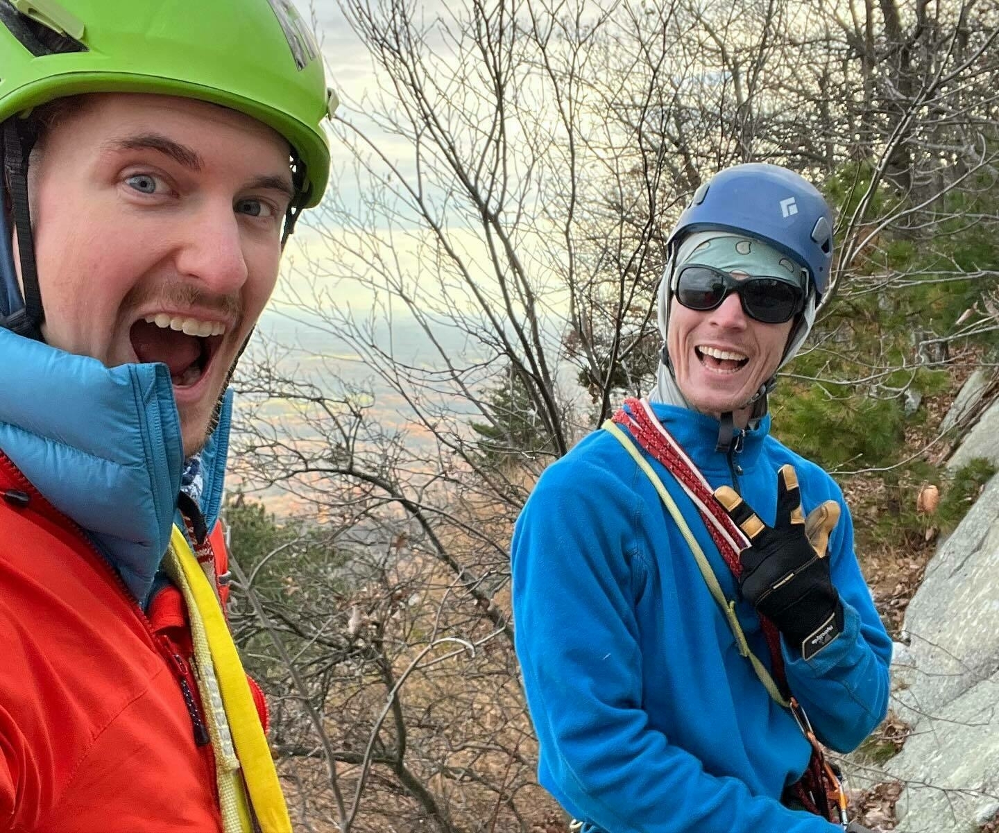 Two climbers wearing helmets are smiling at the camera, showing excitement, with a blurred natural backdrop suggesting an elevated outdoor setting.