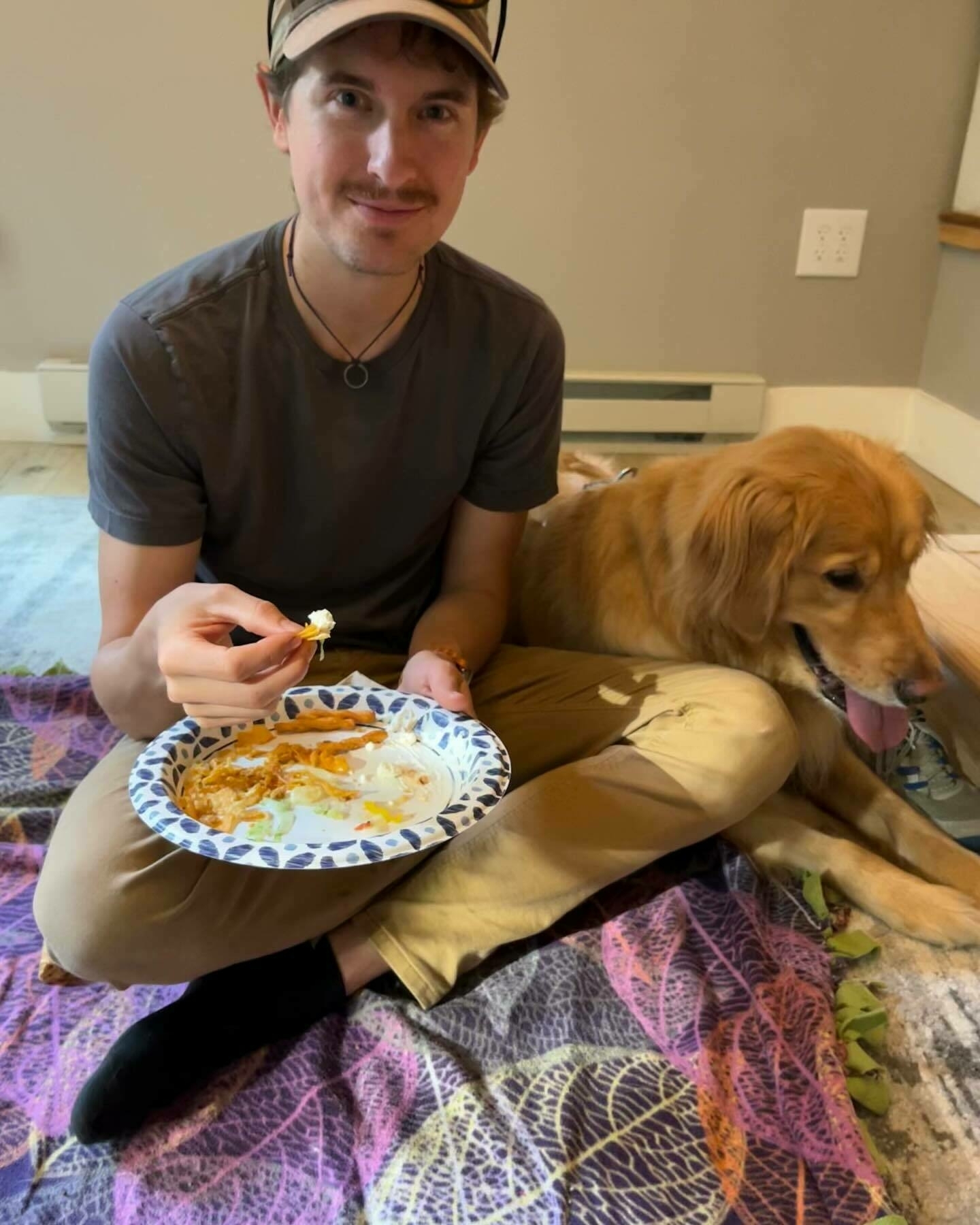 A man sits cross-legged on the floor, eating food, next to a golden retriever, in a casual indoor setting.