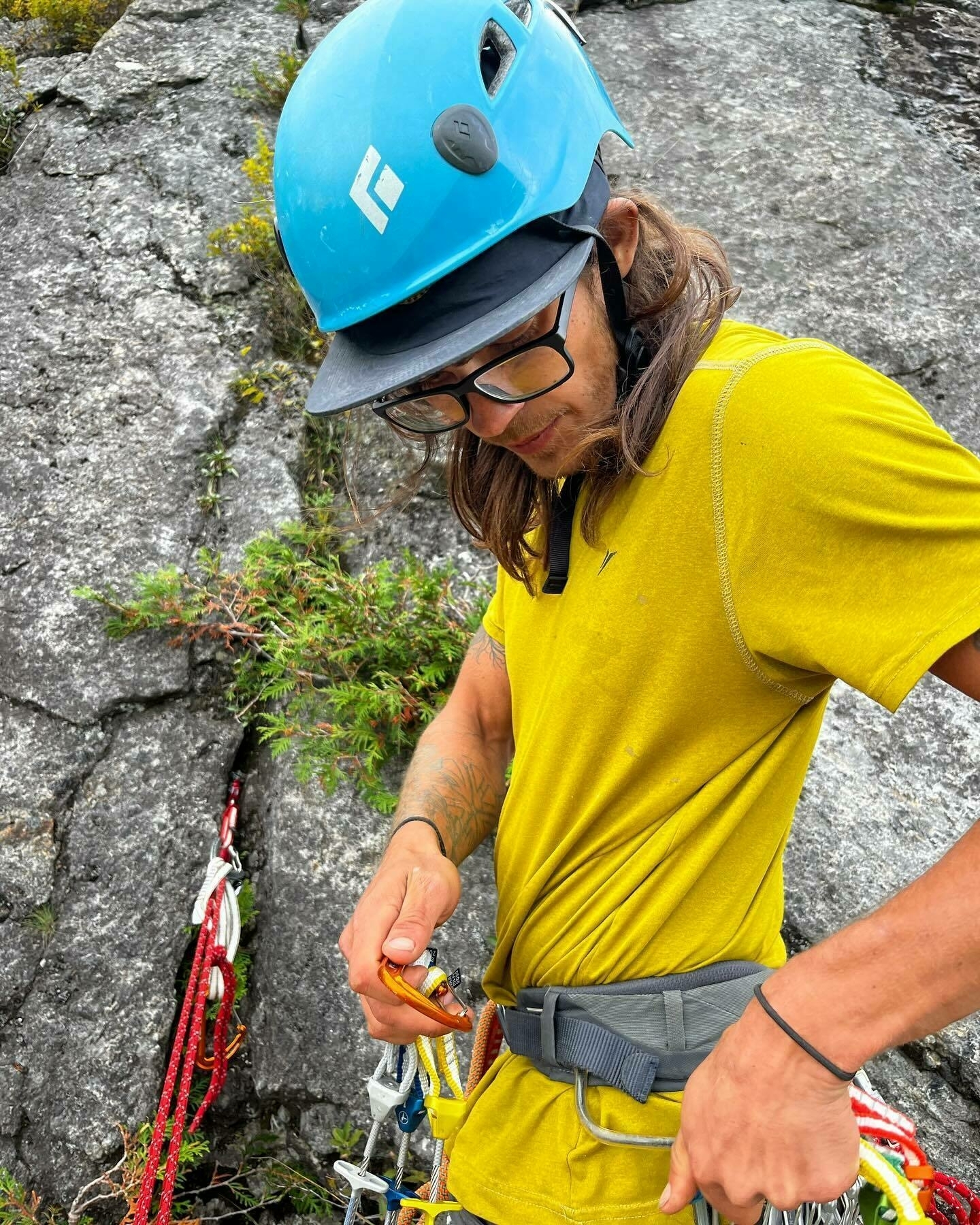 A person in a yellow shirt and blue helmet adjusts climbing equipment against a rock face backdrop.