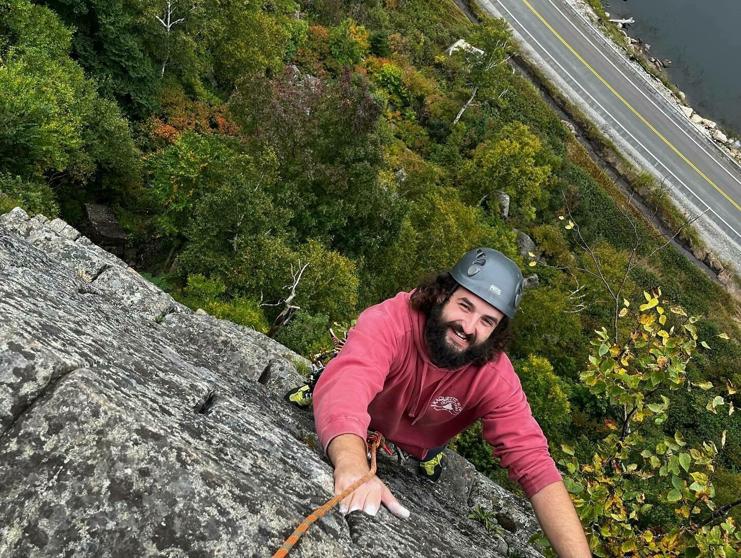 A person in a helmet is rock climbing, smiling at the camera, with a road and dense trees in the background.