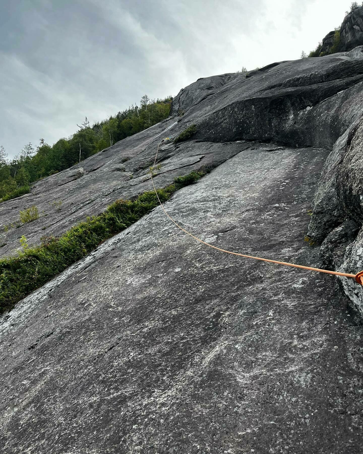A steep, gray rock face with an orange climbing rope snaking up it; surrounded by green foliage under a cloudy sky.