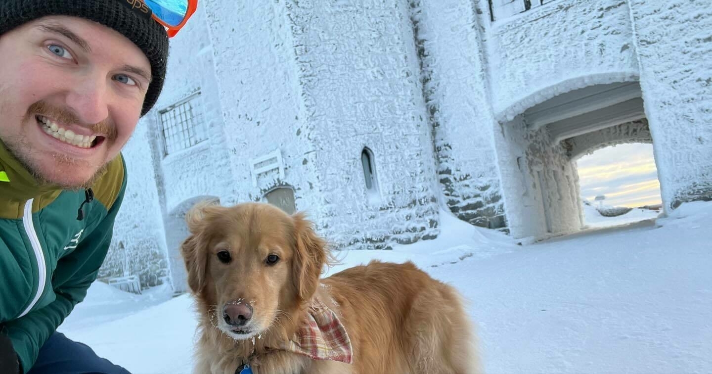 A man and a golden retriever posing together, smiling in a snow-covered landscape with a frosted building in the background.
