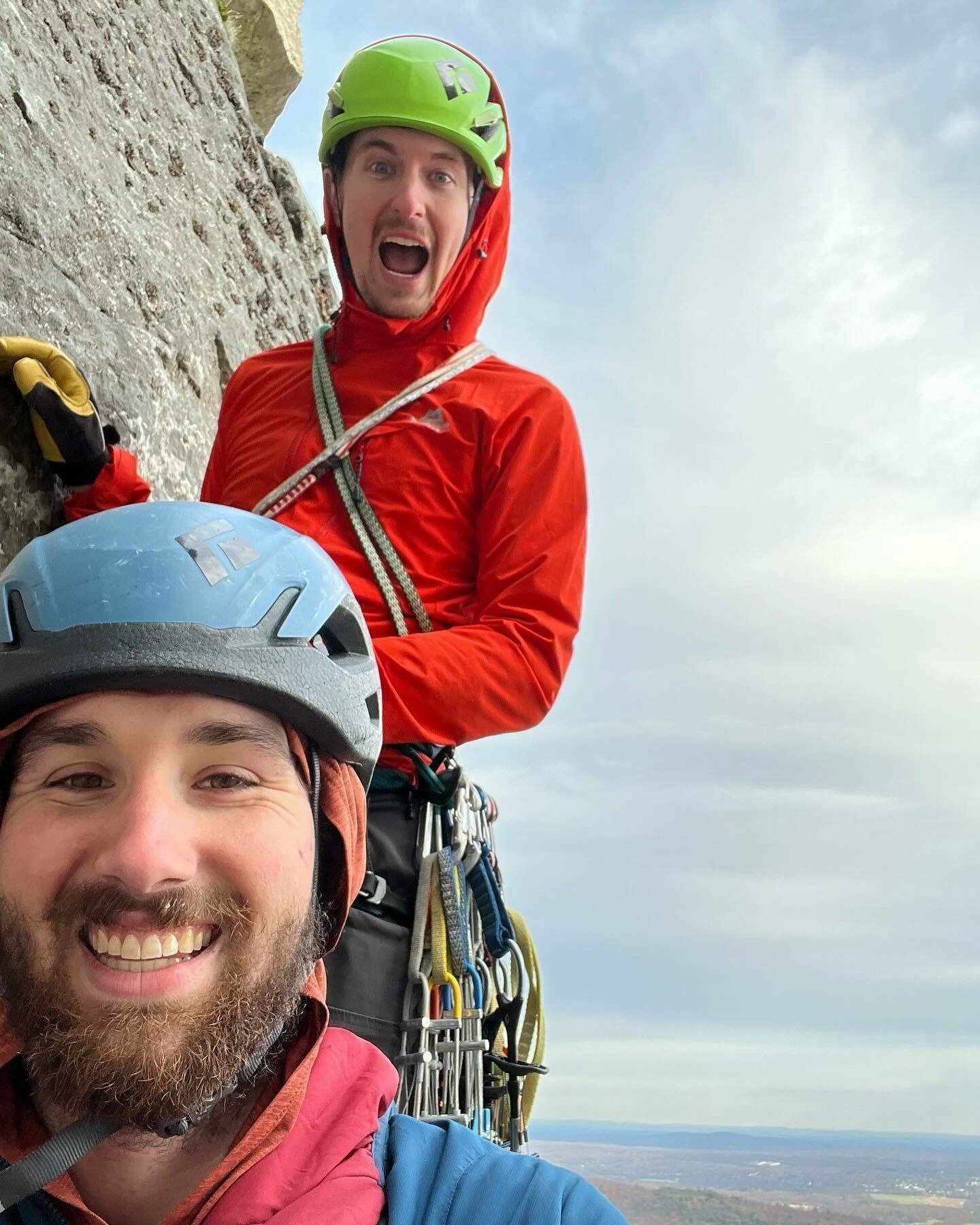 Two climbers in helmets smiling on a rock face; one in foreground is taking the selfie with landscape visible below.