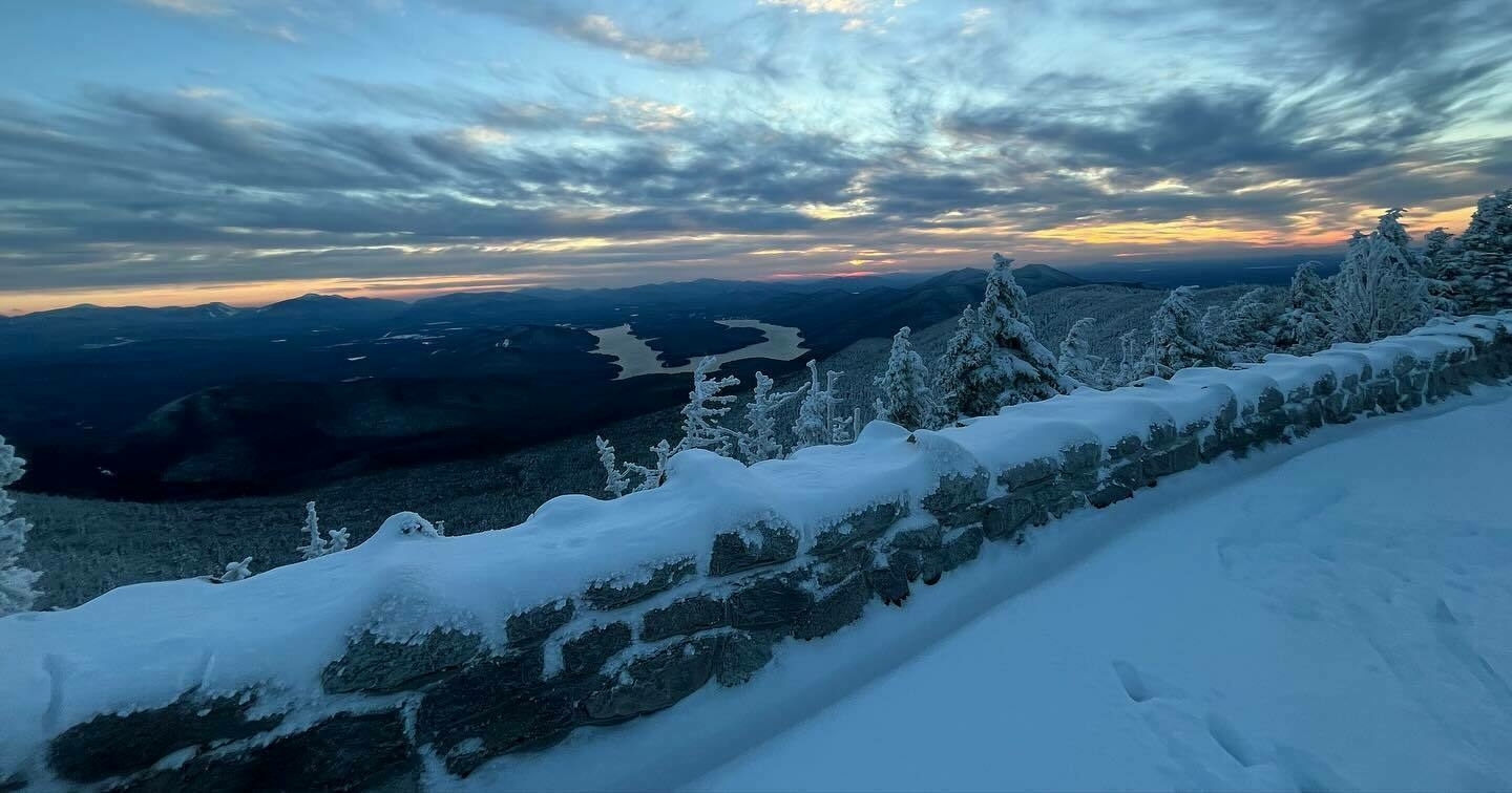 A snowy landscape at dawn with snow-covered trees and a stone wall, overlooking a lake amid mountain ranges under a cloudy sky.