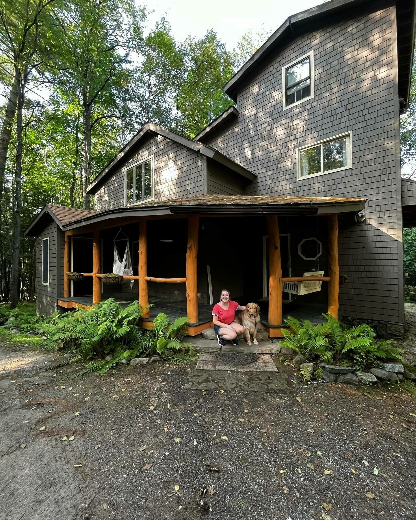 A person seated next to a dog on the porch of a two-story house surrounded by lush greenery.