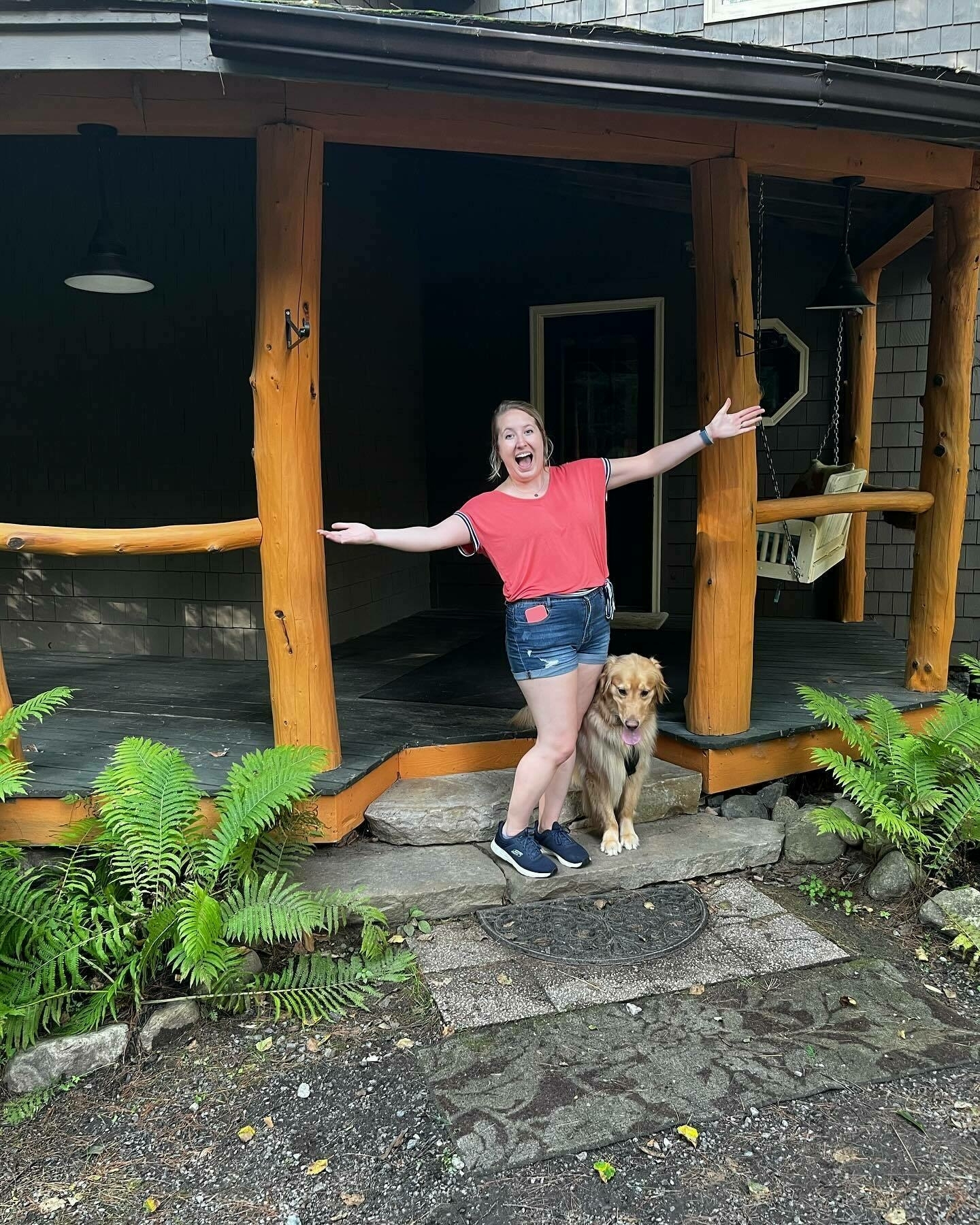 A person excitedly poses with outstretched arms next to a dog on a porch surrounded by ferns.