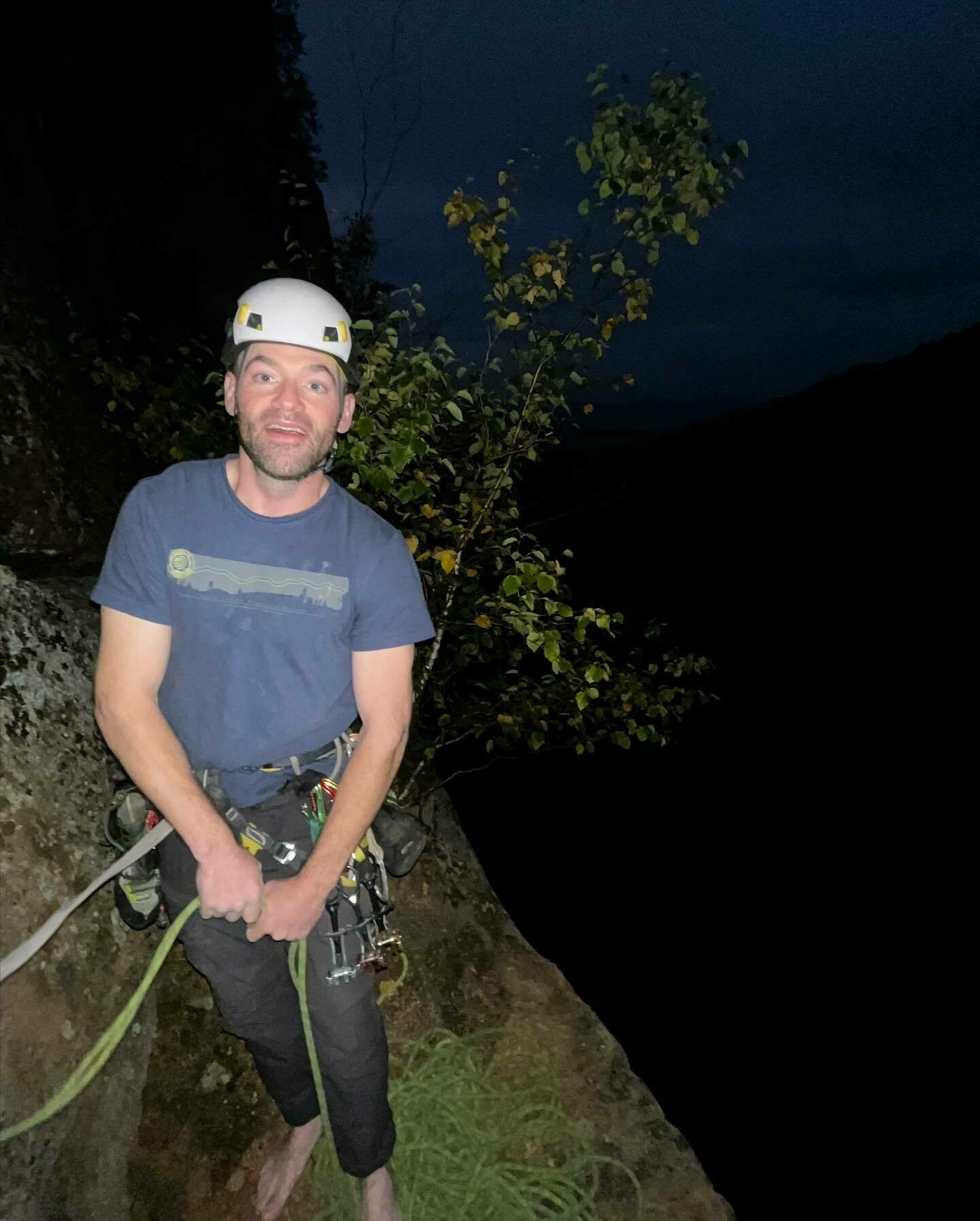 A person in climbing gear stands on a rock ledge at night, smiling, with a dark, natural backdrop.