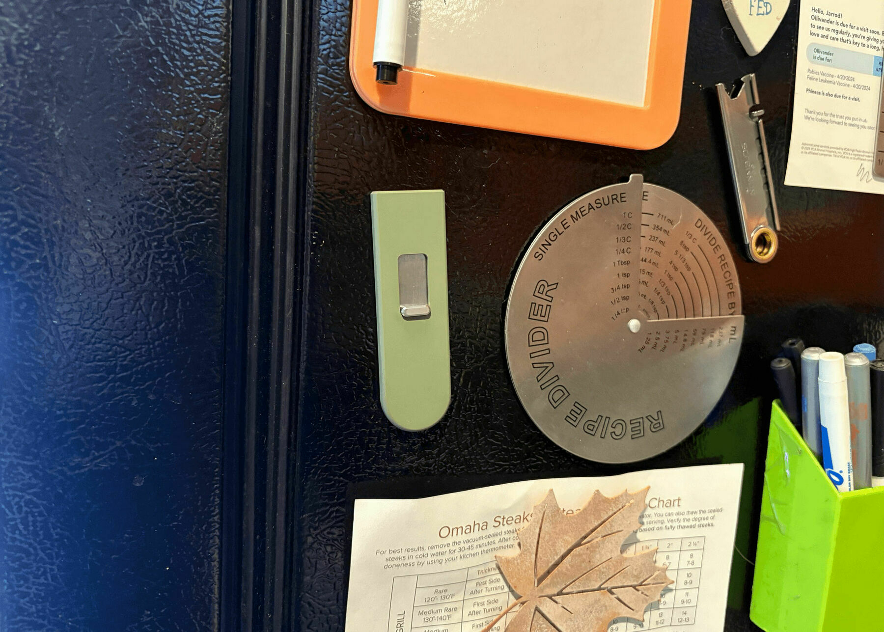 A refrigerator door with several items attached to it. There’s a magnetic dry erase board with a marker, a round metal recipe measurement divider with metric and teaspoon conversions, and a green Keen utility knife. There’s also a piece of paper with steak cooking instructions, secured by a leaf-shaped magnet. A green holder contains a few dry erase markers on the right side.