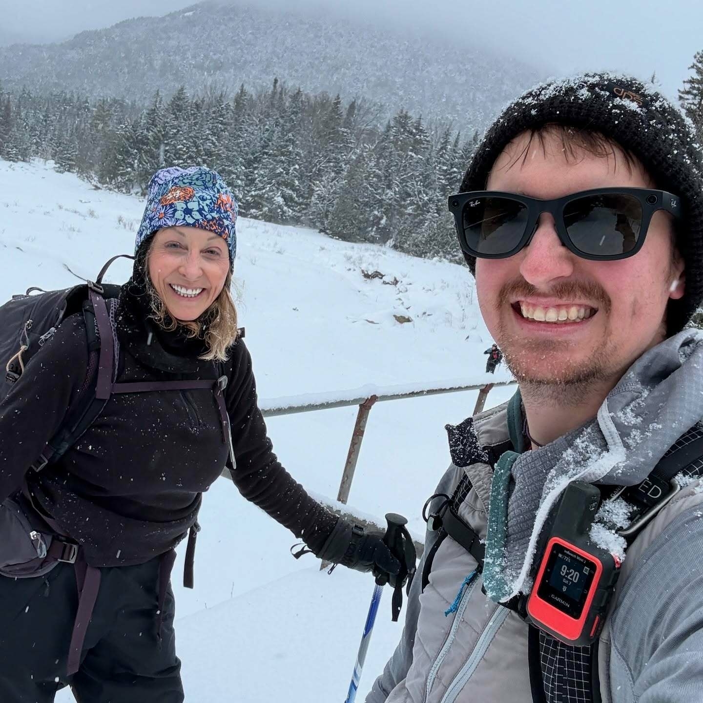 Two people smiling stand in a snowy landscape holding ski poles surrounded by snow-covered trees and mountains wearing winter clothing and backpacks. A device on the person’s backpack reads 9:20.