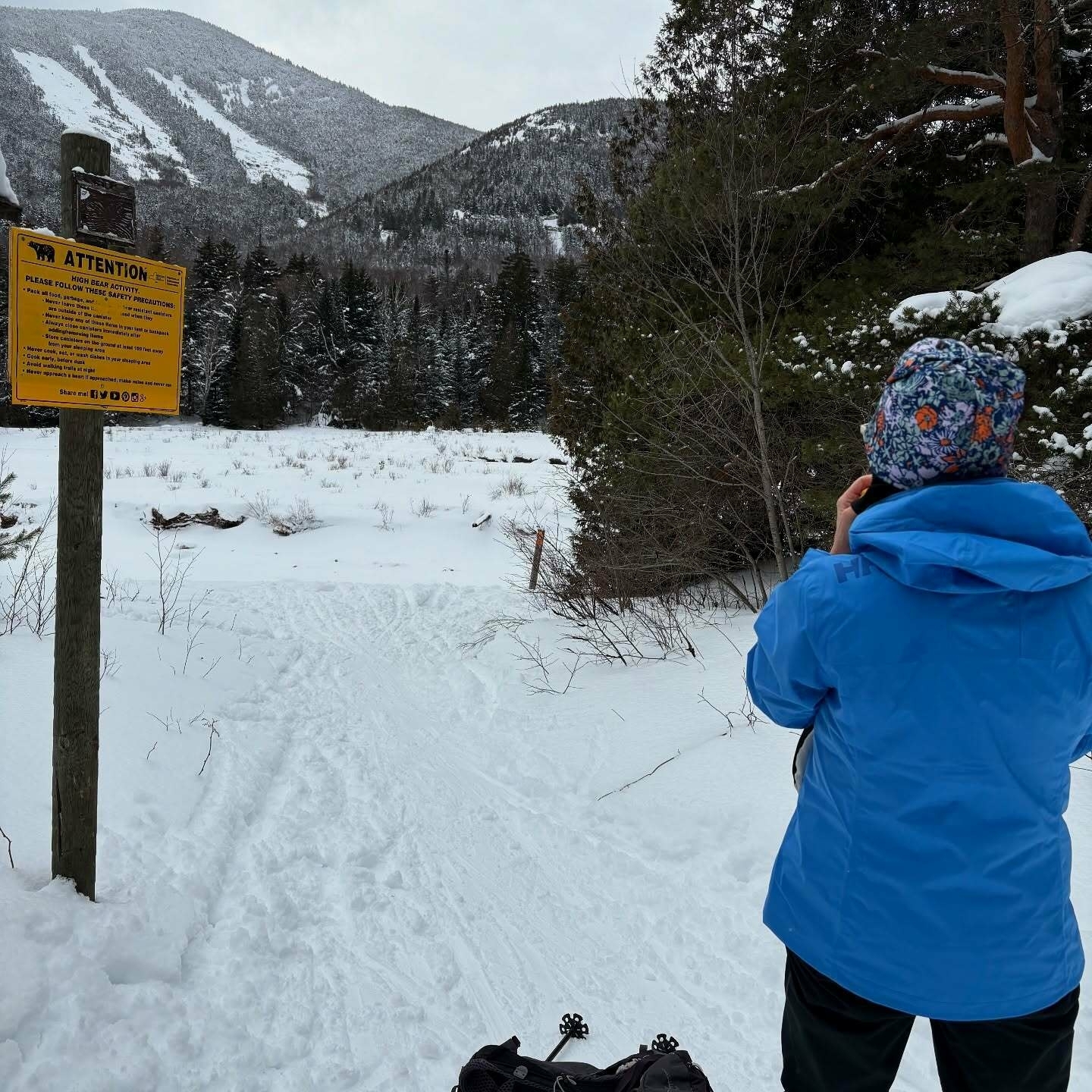 A person in a blue jacket faces snow-covered mountains, holding a camera. A wooden post with a yellow sign reads: ATTENTION Please move these objects and follow these safety instructions. (Illegible text).