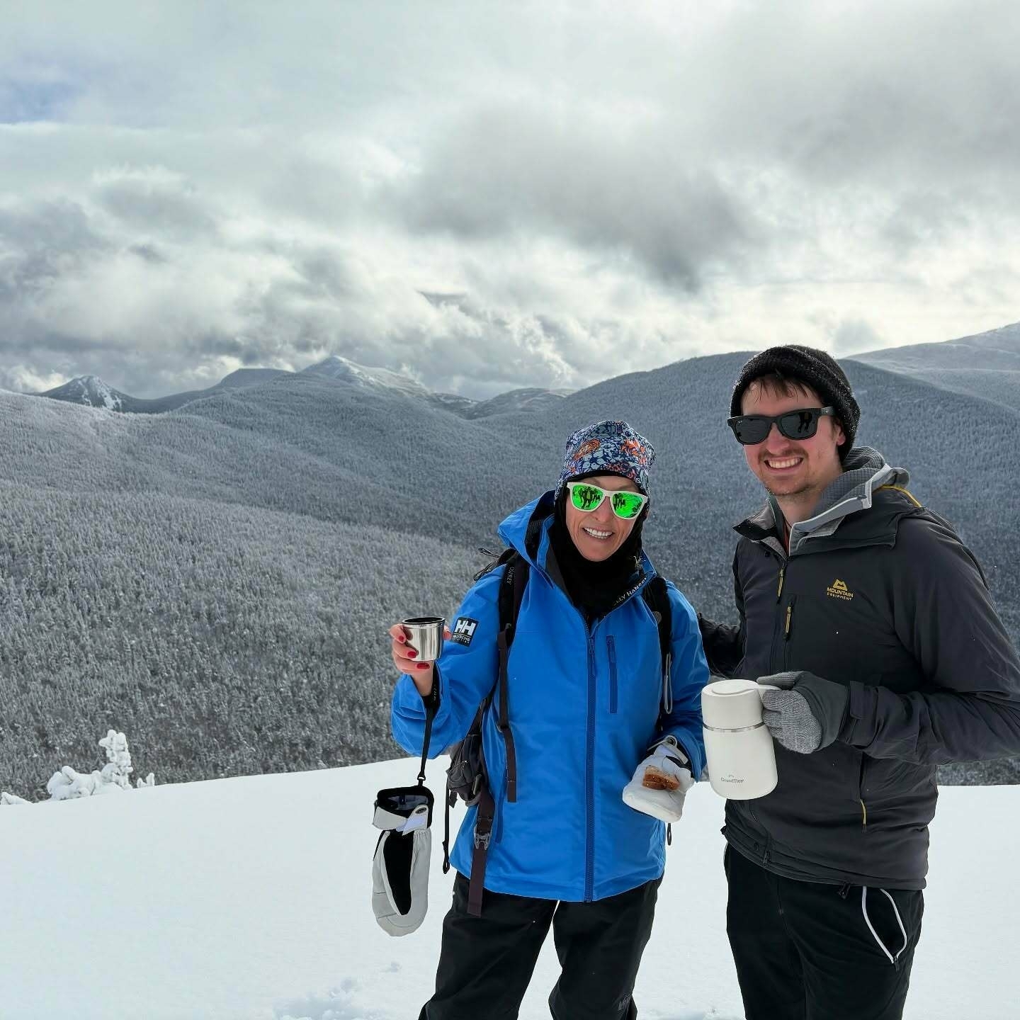 Two people wearing winter clothing hold mugs on a snowy mountaintop with forested mountains and cloudy skies in the background.