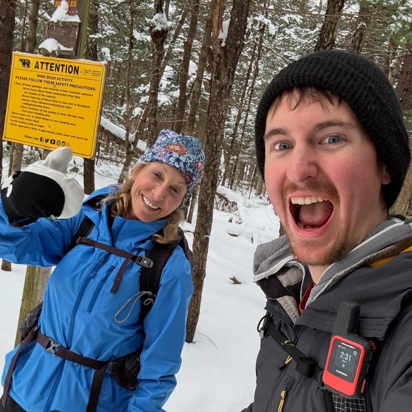 Two smiling people in winter clothing stand in a snowy forest. A yellow sign behind them reads ATTENTION HIGH BEAR ACTIVITY PLEASE FOLLOW THESE SAFETY PRECAUTIONS and lists several safety tips.
