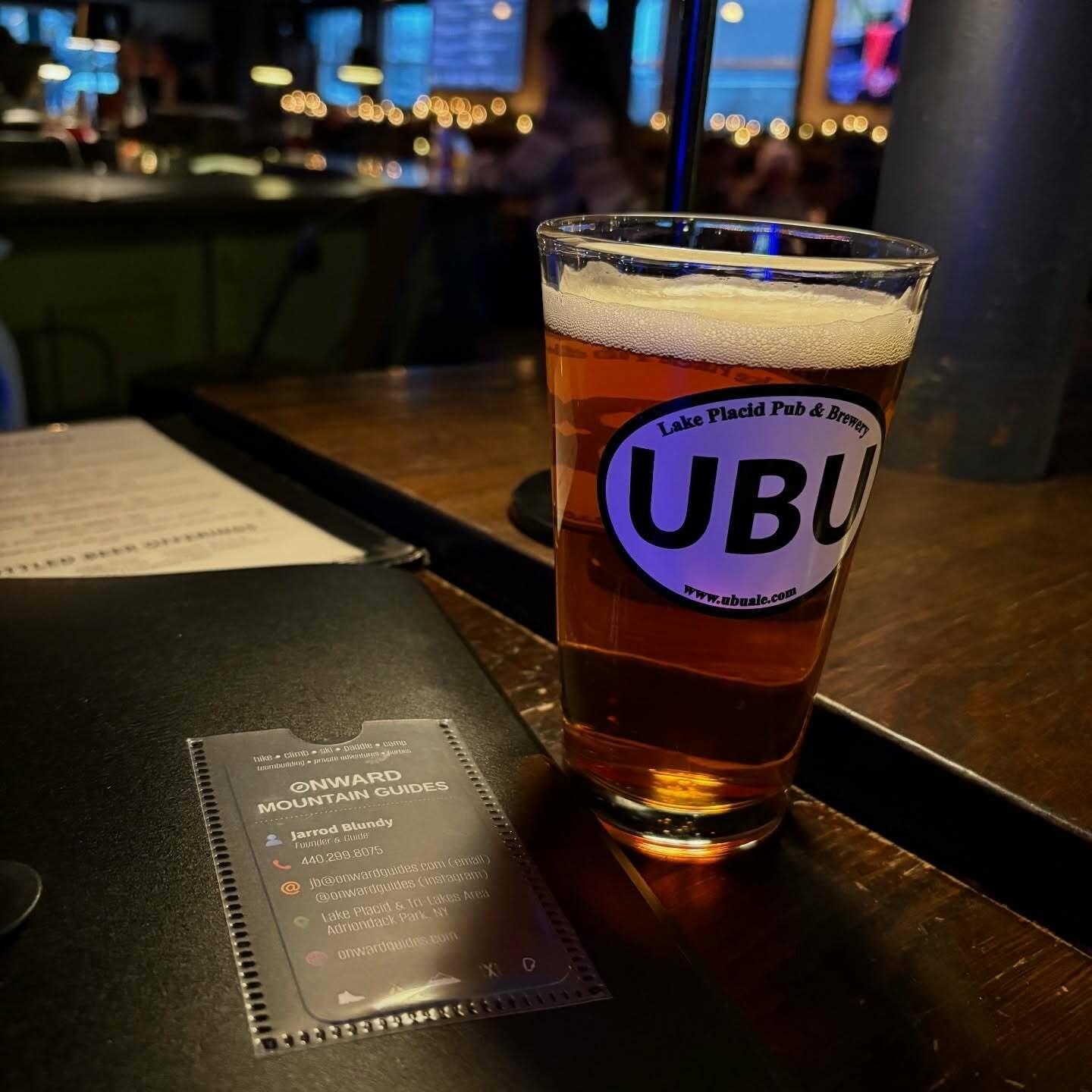A pint glass of amber beer rests on a dimly lit bar counter displaying a &ldquo;UBU Lake Placid Pub & Brewery&rdquo; label. Nearby lies a booklet labeled &ldquo;ONWARD MOUNTAIN GUIDES&rdquo; with contact information for Jarrod Blundy, including phone number 440-289-8075 and email onwardmountainguides@gmail.com. The guide shop is located in Lake Placid and the High Peaks Area of the Adirondack Park NY. Various symbols and social media handles are shown.