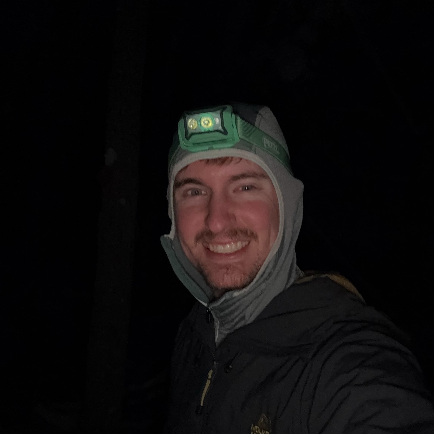 A person wearing a headlamp and a hoodie smiles in darkness. The headlamp is green with the brand name Petzl illuminated, suggesting nighttime or low-light conditions outdoors.