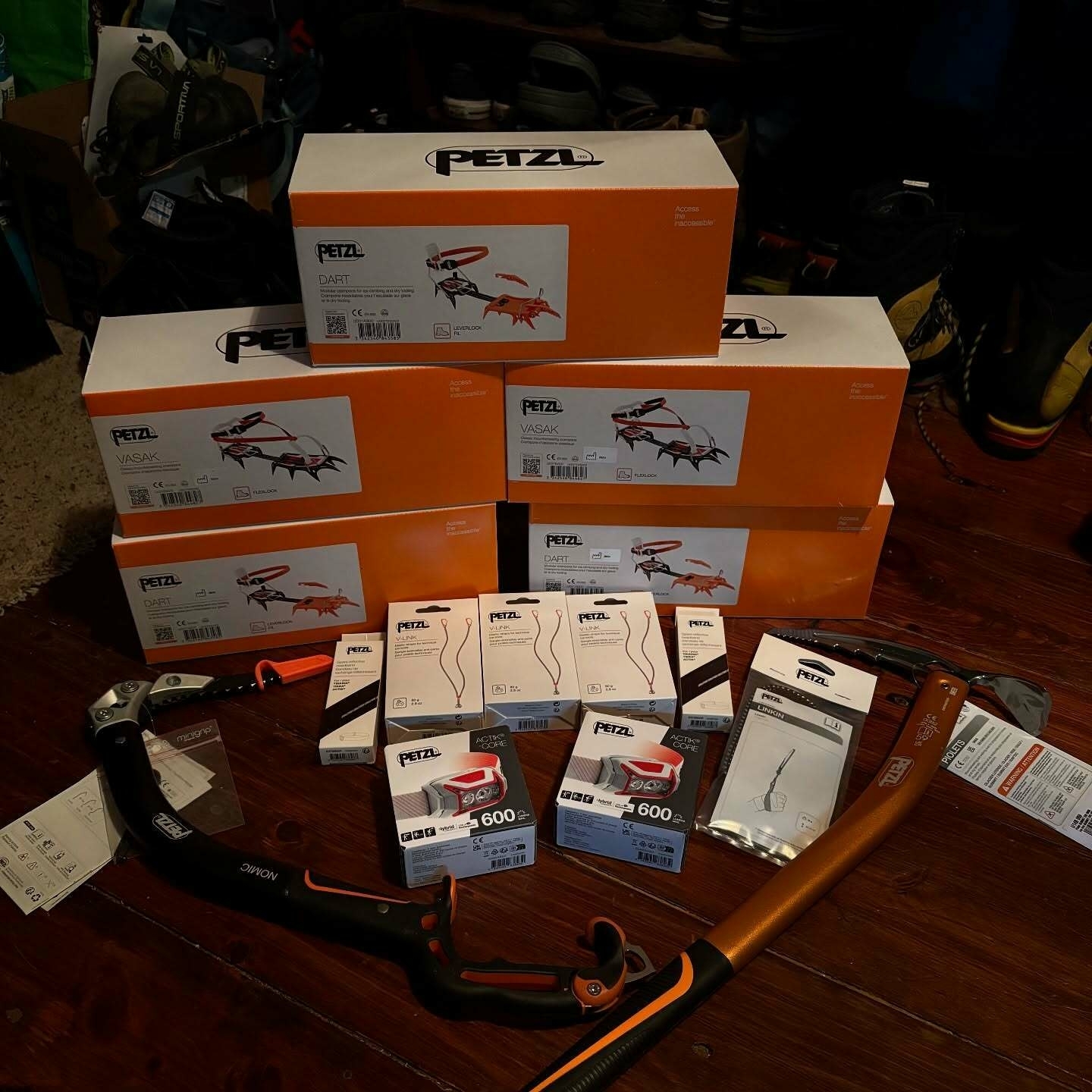 Boxes and climbing equipment are stacked on a wooden floor. The orange and white boxes are from Petzl, labeled &ldquo;Dart&rdquo; and &ldquo;Vasak.&rdquo; Climbing gear is displayed around them, including ice axes.