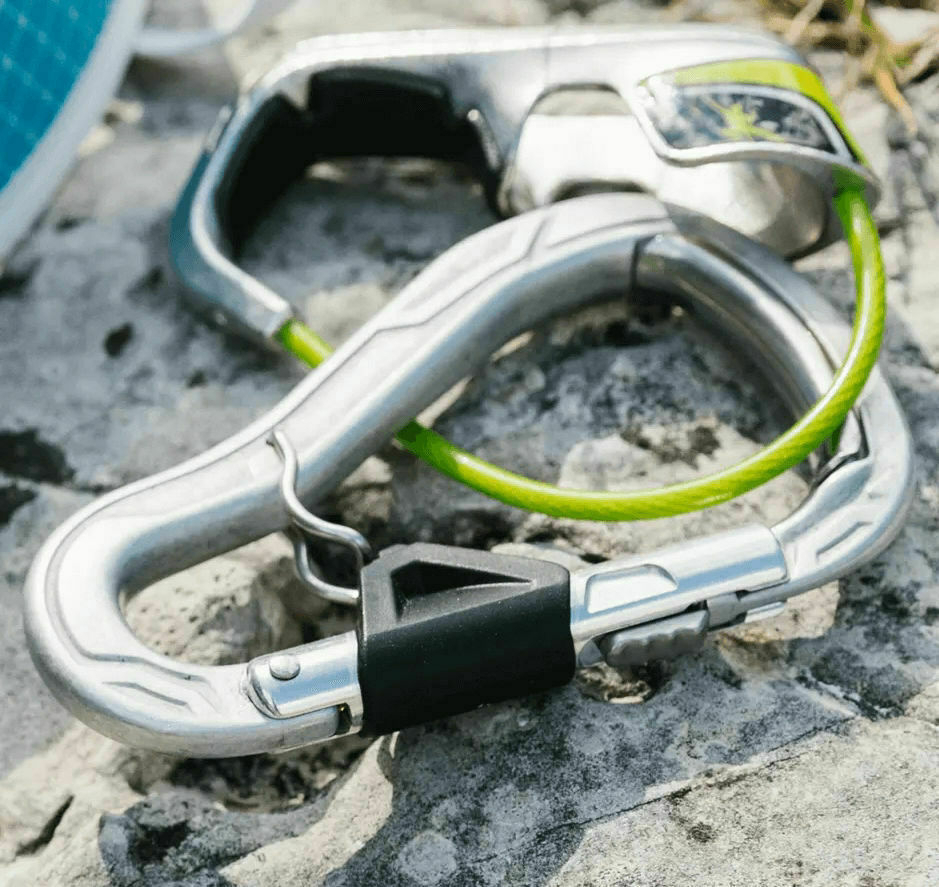Close up of a gray carabiner attached to a belay device upon a rocky surface.